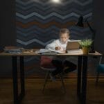 Chłopiec odrabia lekcje przy biurku z tabletem, lampką i książkami