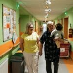 Dwie kobiety idą korytarzem domu opieki, jedna trzyma lalkę dziecka